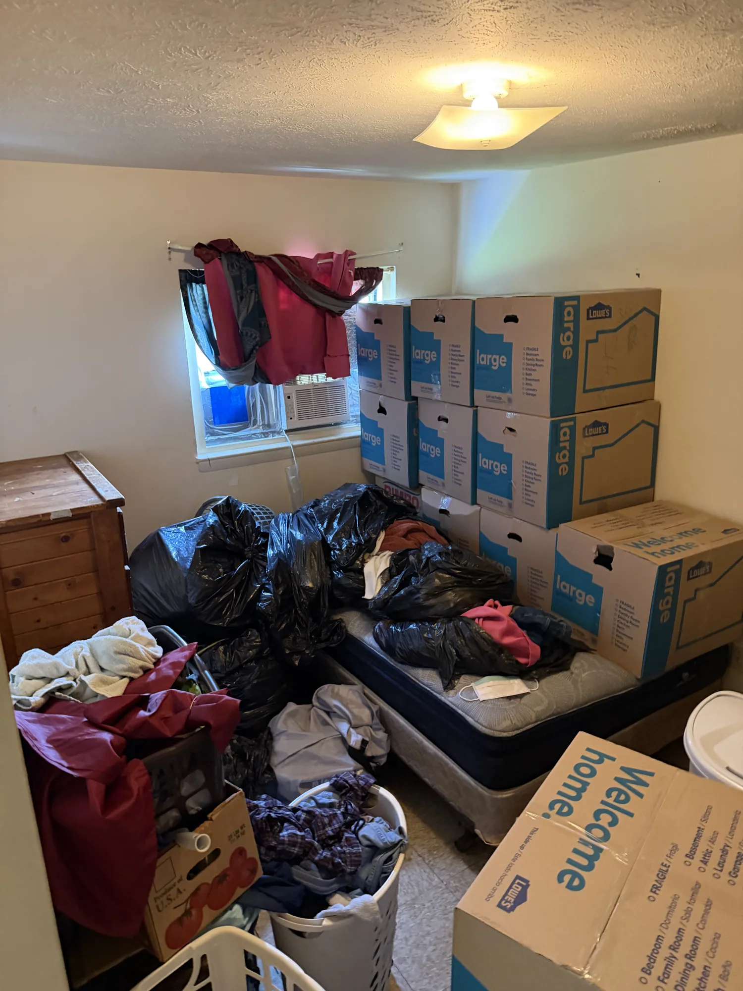 Housing Authority bedroom before cleanout, stacked with large moving boxes, bags of clothes and laundry baskets by a red-curtained window.