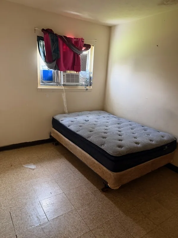 The same bedroom cleared out, red-curtained window and AC unit, bare mattress on the frame and empty tile floor.